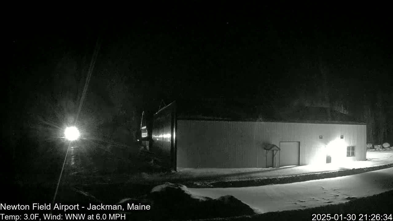 A long, low building is illuminated at night in snowy conditions with a 3°F temperature and a 6.0 mph wind from the west-northwest.