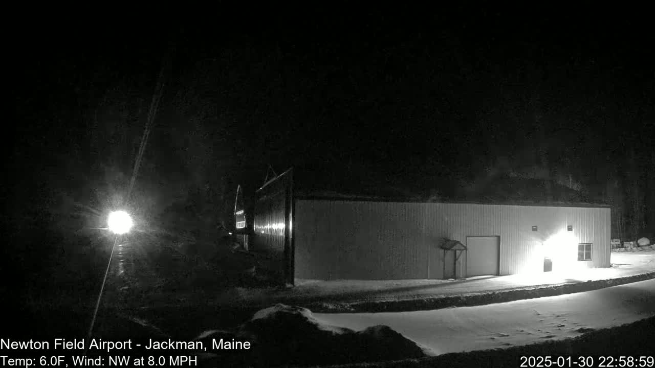 A long, low building is illuminated at night in snowy conditions with a northwest wind.