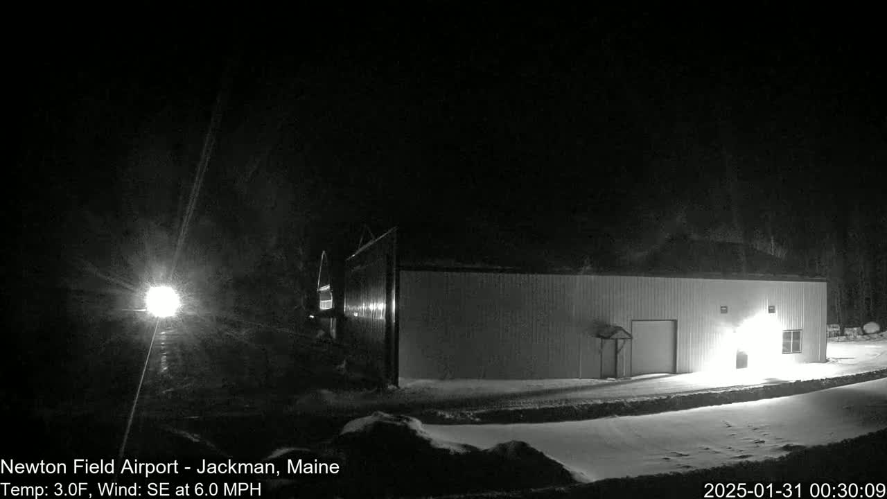A snow-covered, brightly lit hangar sits in the foreground under a dark, nighttime sky with a southeast wind.