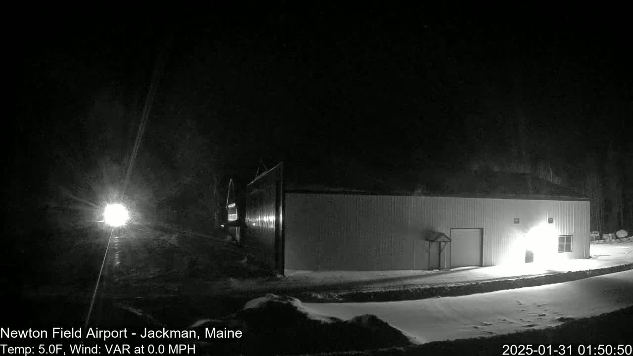 A long, low building is illuminated at night in the snow, with a vehicle's headlights visible in the distance.