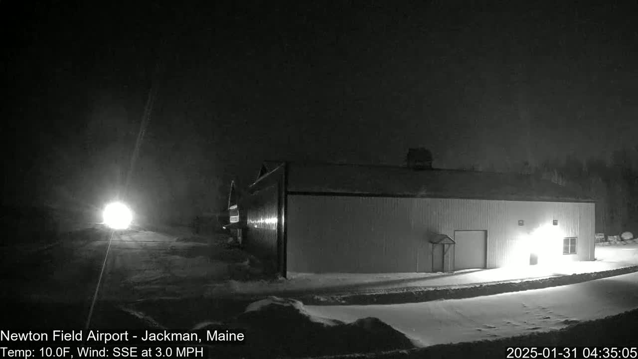 A long, low building is illuminated at night in snowy conditions, with a bright light visible in the distance.