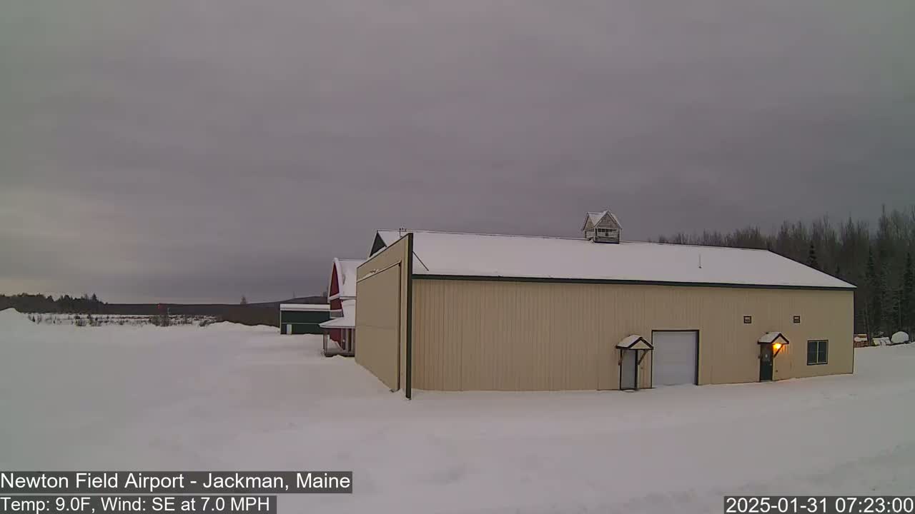 A tan hangar with a snow-covered roof stands in a snow-covered field under an overcast sky.