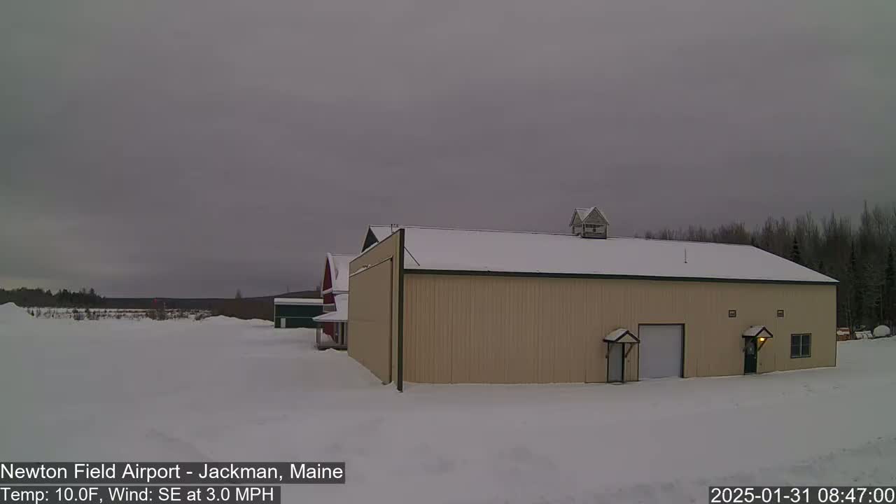 A tan hangar and a red barn sit in a snow-covered field under an overcast sky.