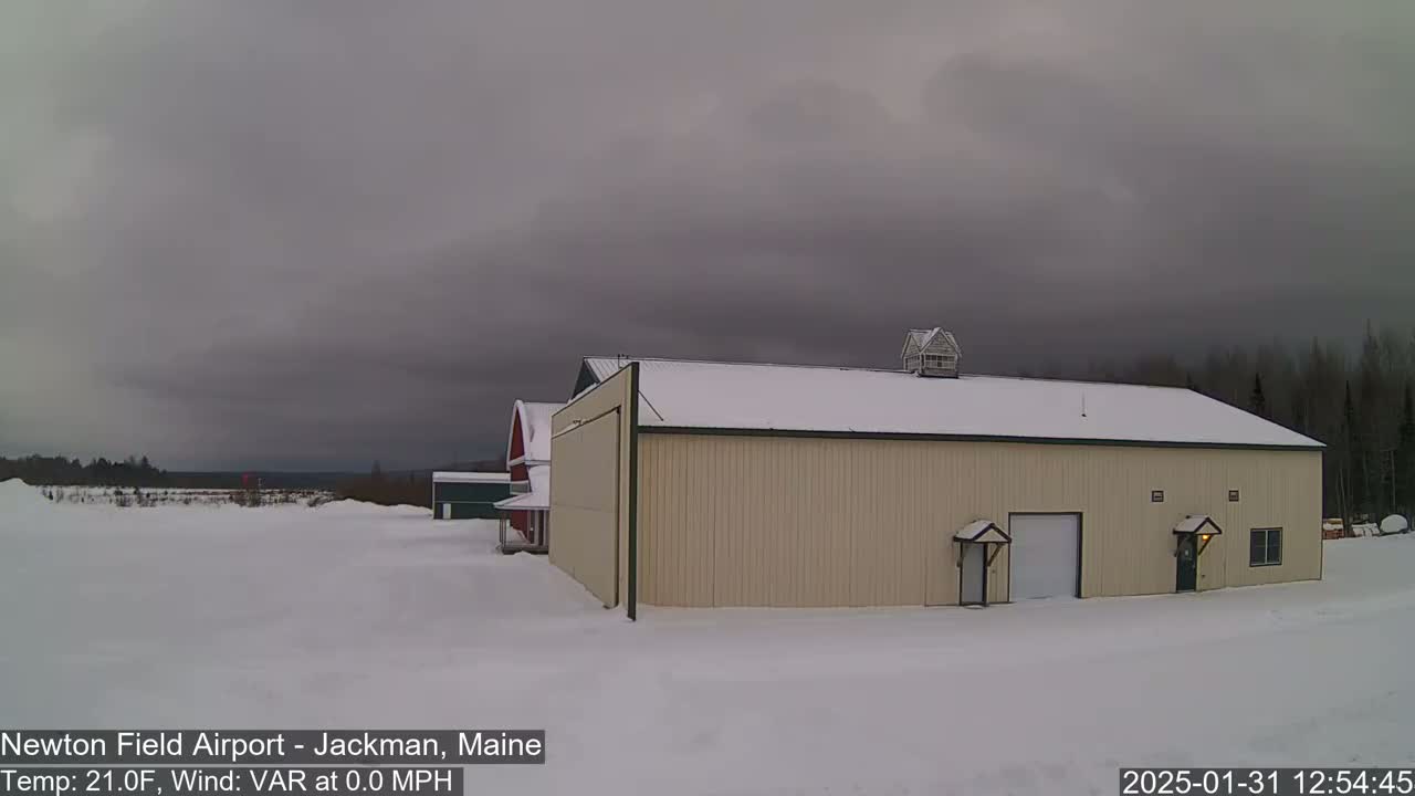 A tan hangar with a snow-covered roof sits in a snowy landscape under an overcast sky.