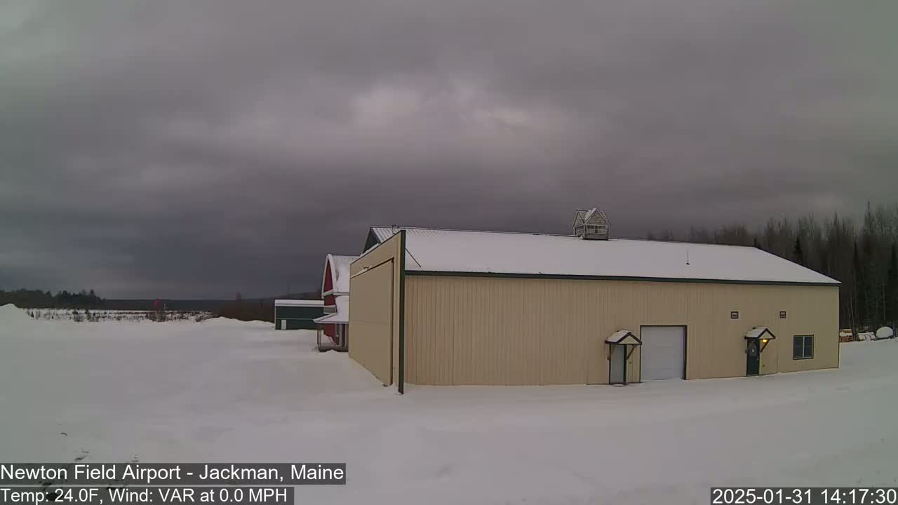A large tan hangar and a smaller red barn sit on a snow-covered field under an overcast sky.