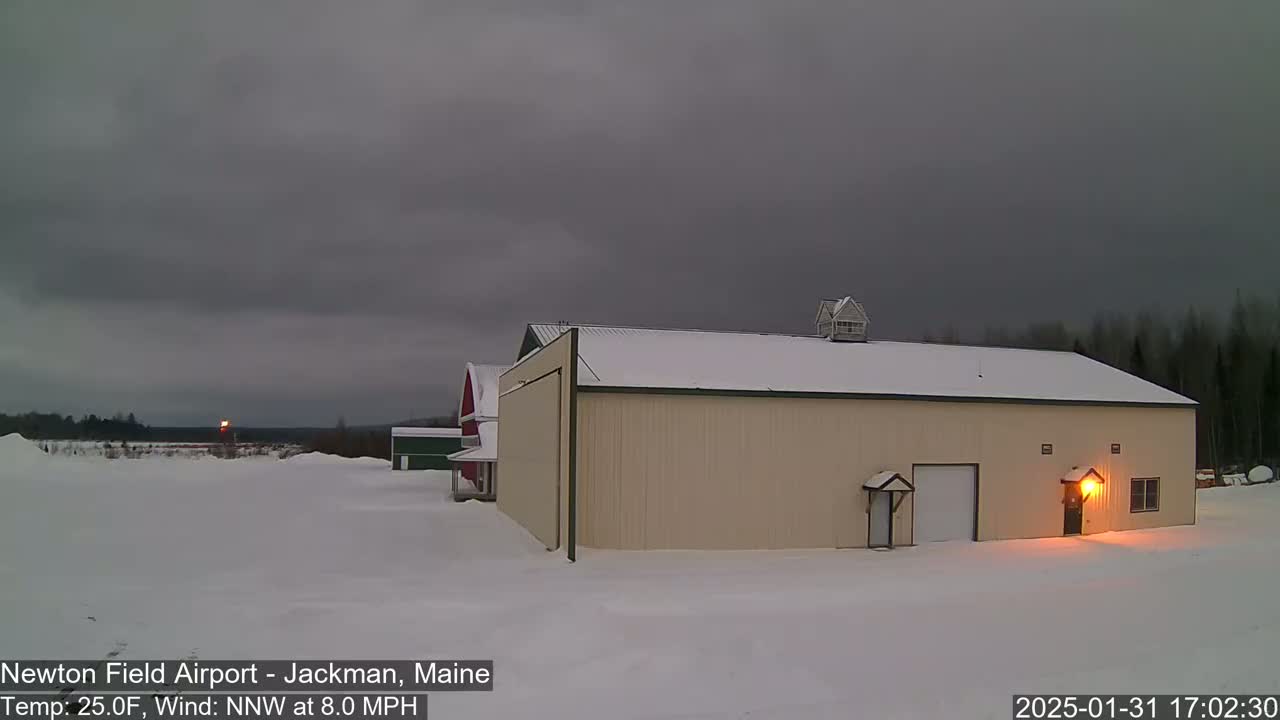 A large tan hangar sits in a snow-covered field under an overcast sky, with a smaller red barn visible in the background.