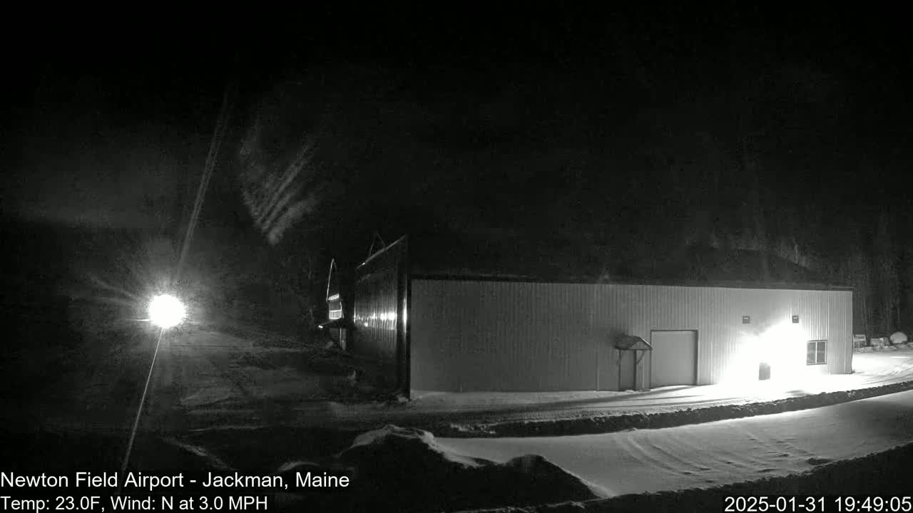 A long, low building sits in a snowy landscape at night under a dark sky with light snow blowing.