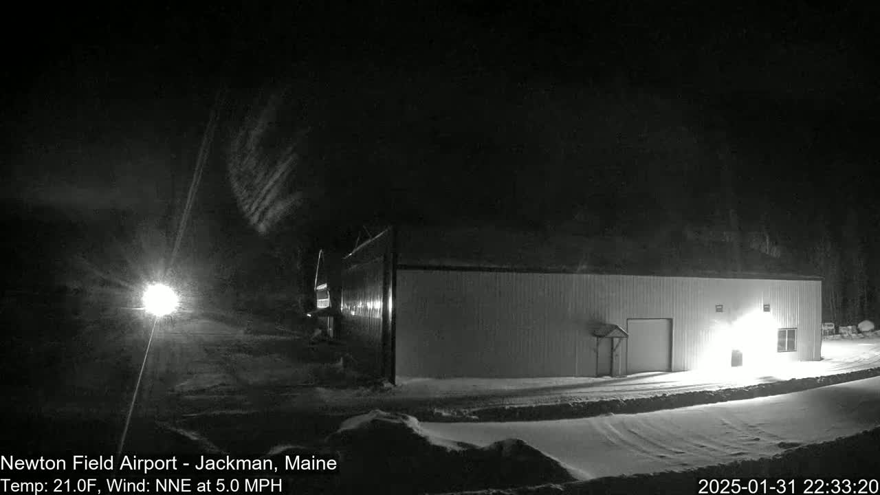 A snow-covered landscape at night shows a long, low building illuminated by lights, under a dark sky with faint clouds and a light wind.