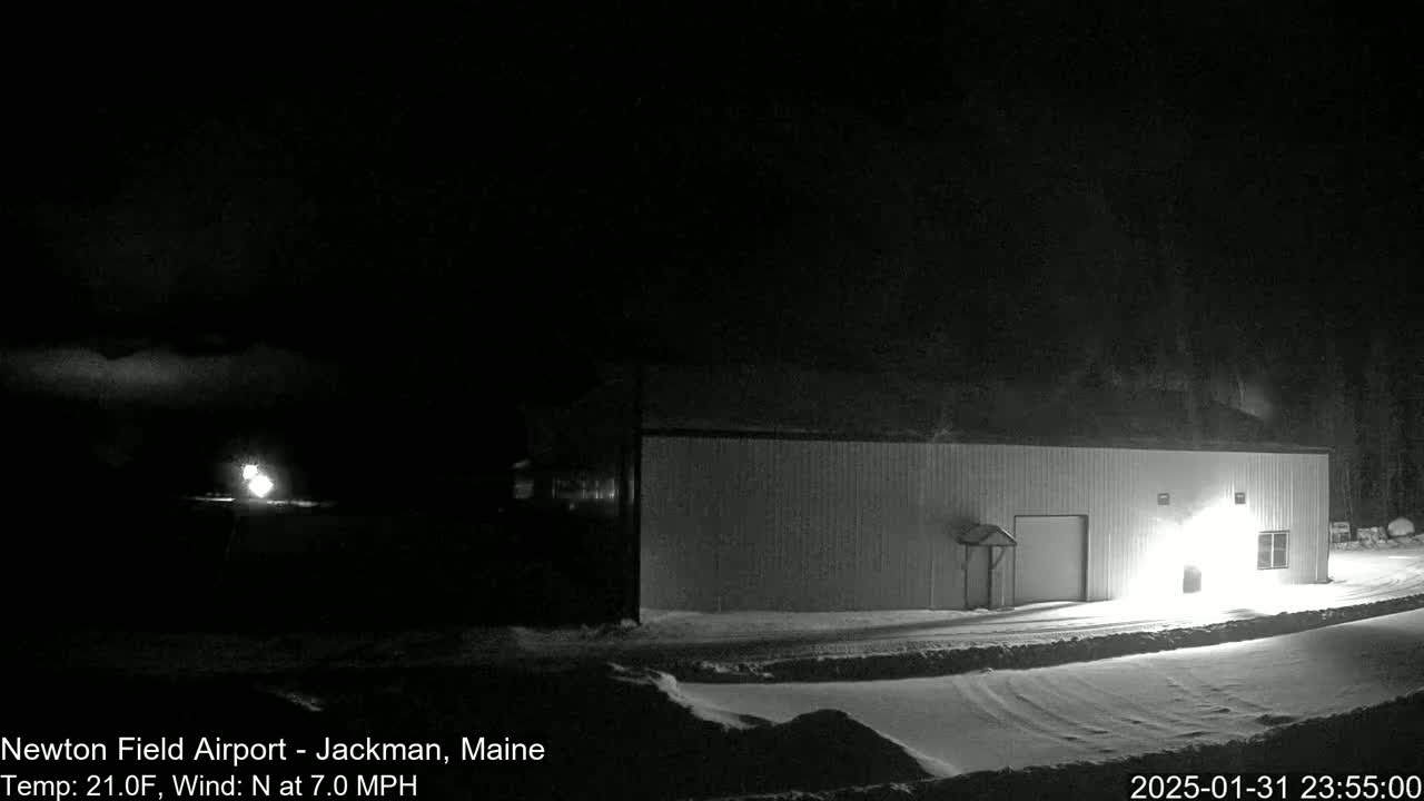 A long, light-colored building is visible at night in snowy conditions with a 7 mph northerly wind and a temperature of 21°F.