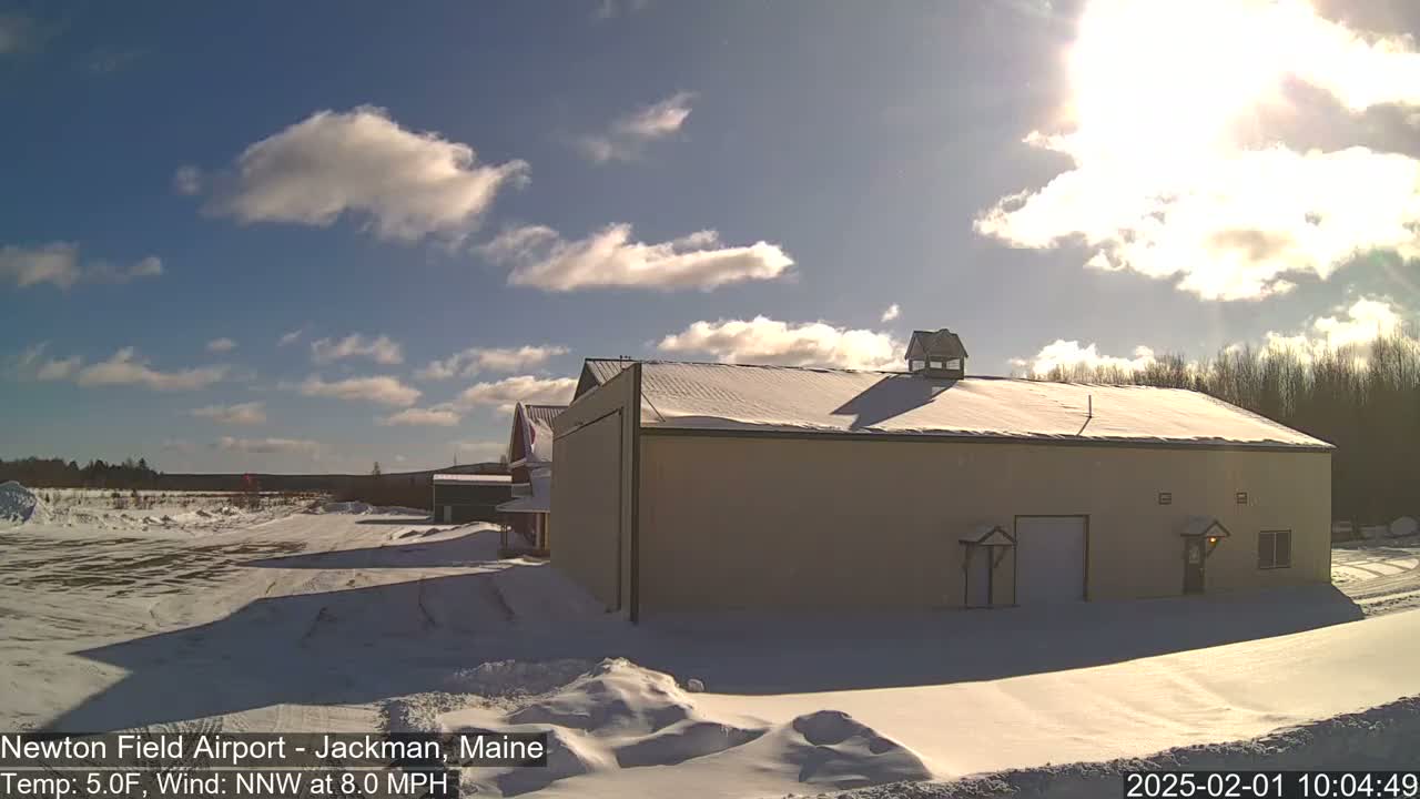A snow-covered airfield with a large tan building under a partly sunny sky.