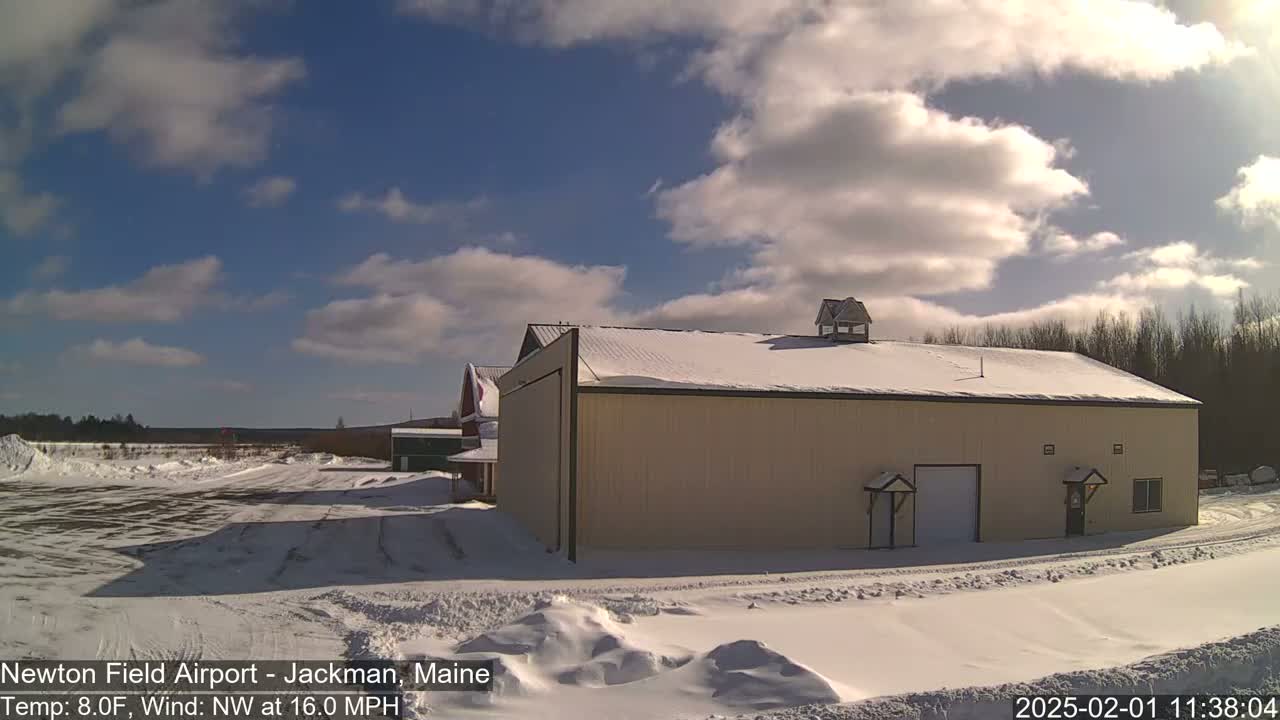 A snow-covered airfield with a large tan building under a partly cloudy, sunny sky.