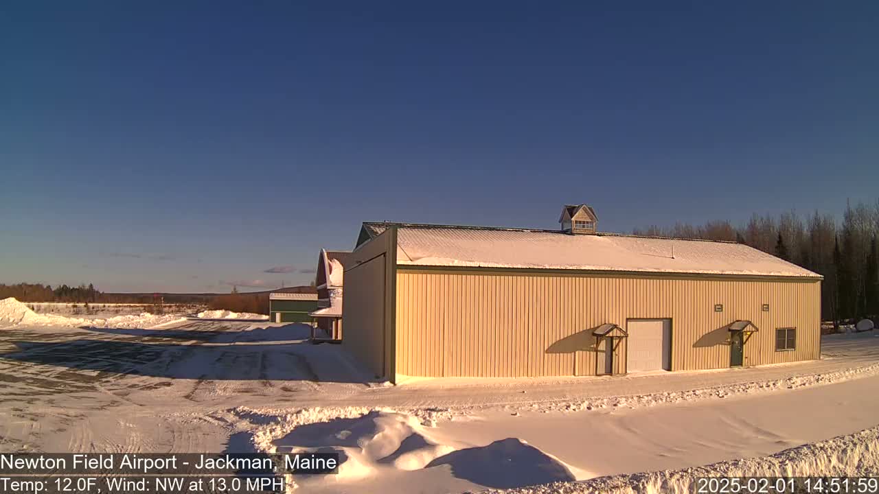 A large tan hangar sits in a snow-covered airfield under a clear blue sky, with a light wind blowing.