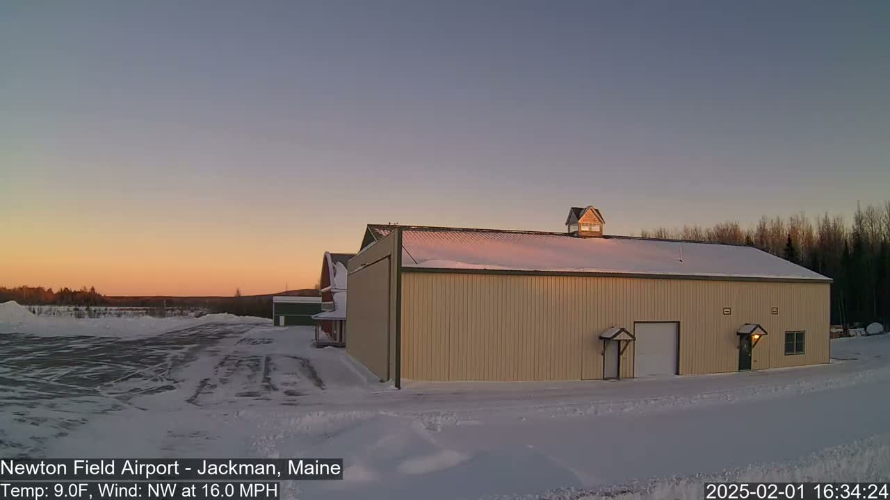 A large tan hangar sits in a snow-covered field at sunset under a clear sky with a northwest wind.