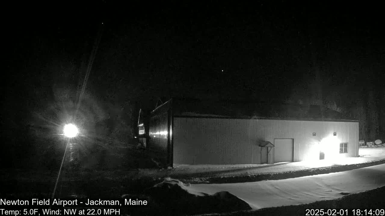 A long, low building is illuminated at night in snowy conditions with a northwest wind at 22 mph and a temperature of 5 degrees Fahrenheit.