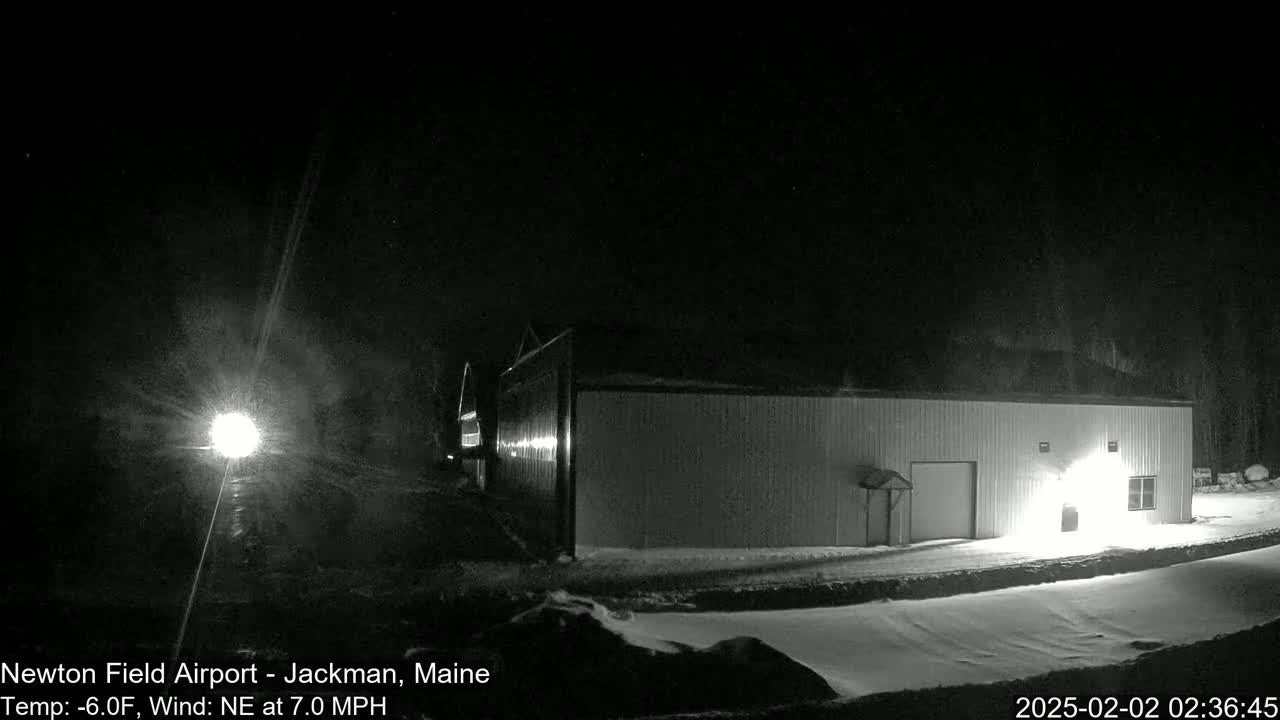 A long, low building is illuminated at night in snowy conditions with a northeast wind of 7 mph and a temperature of -6°F.
