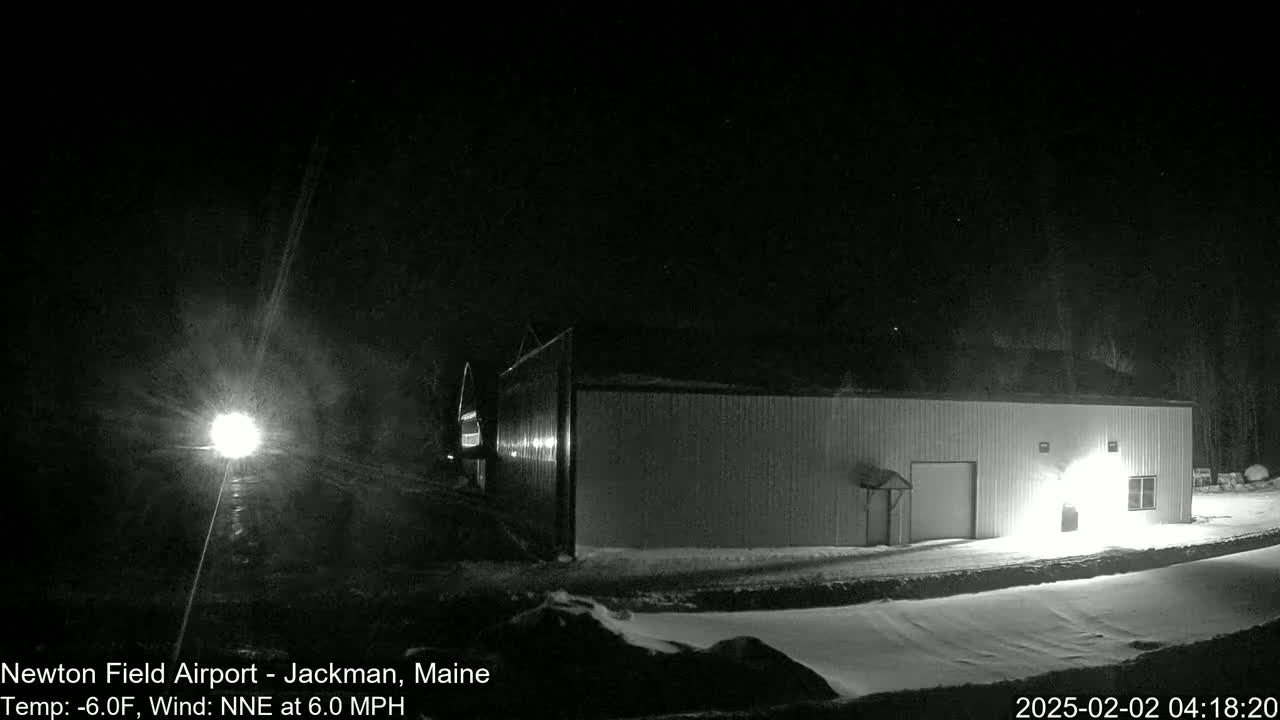 A long, low building is illuminated at night in snowy conditions, with a bright light visible in the distance.