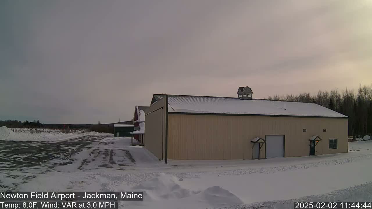 A large tan hangar sits in a snow-covered airfield under an overcast sky.