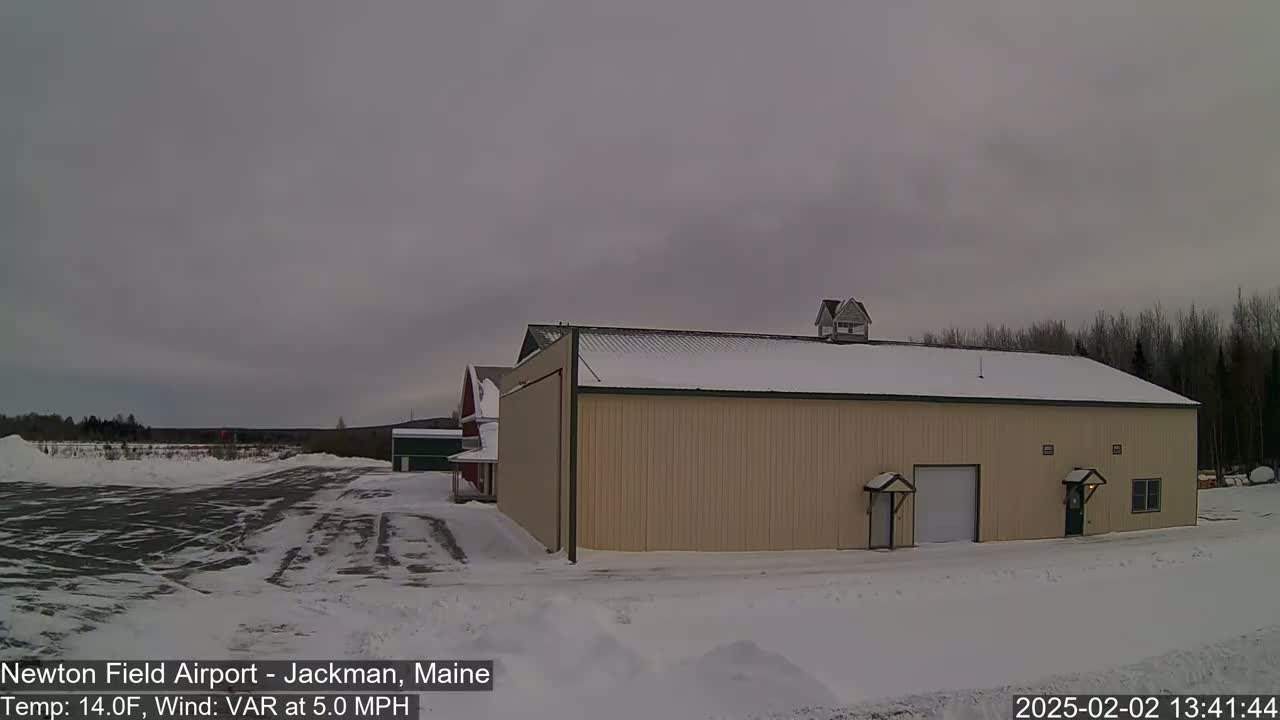 A large tan hangar sits in a snow-covered airfield under an overcast sky.