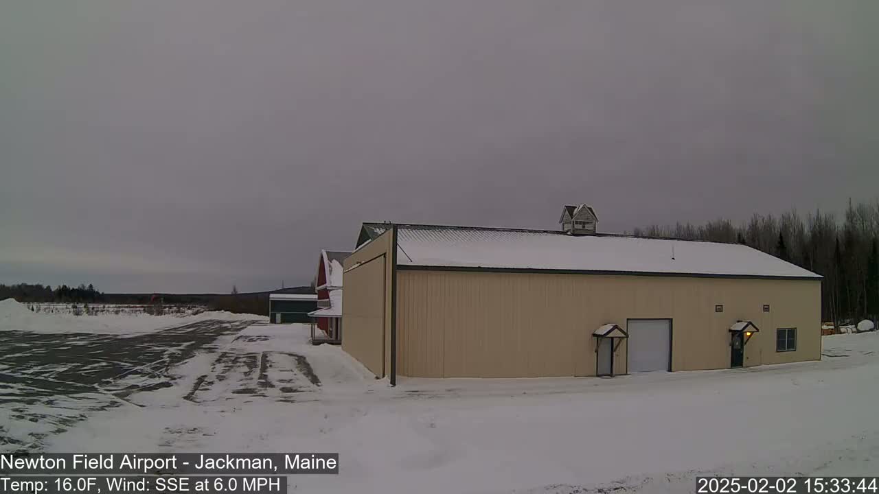 A large tan building with a snow-covered roof stands in a snow-covered airfield under an overcast sky.