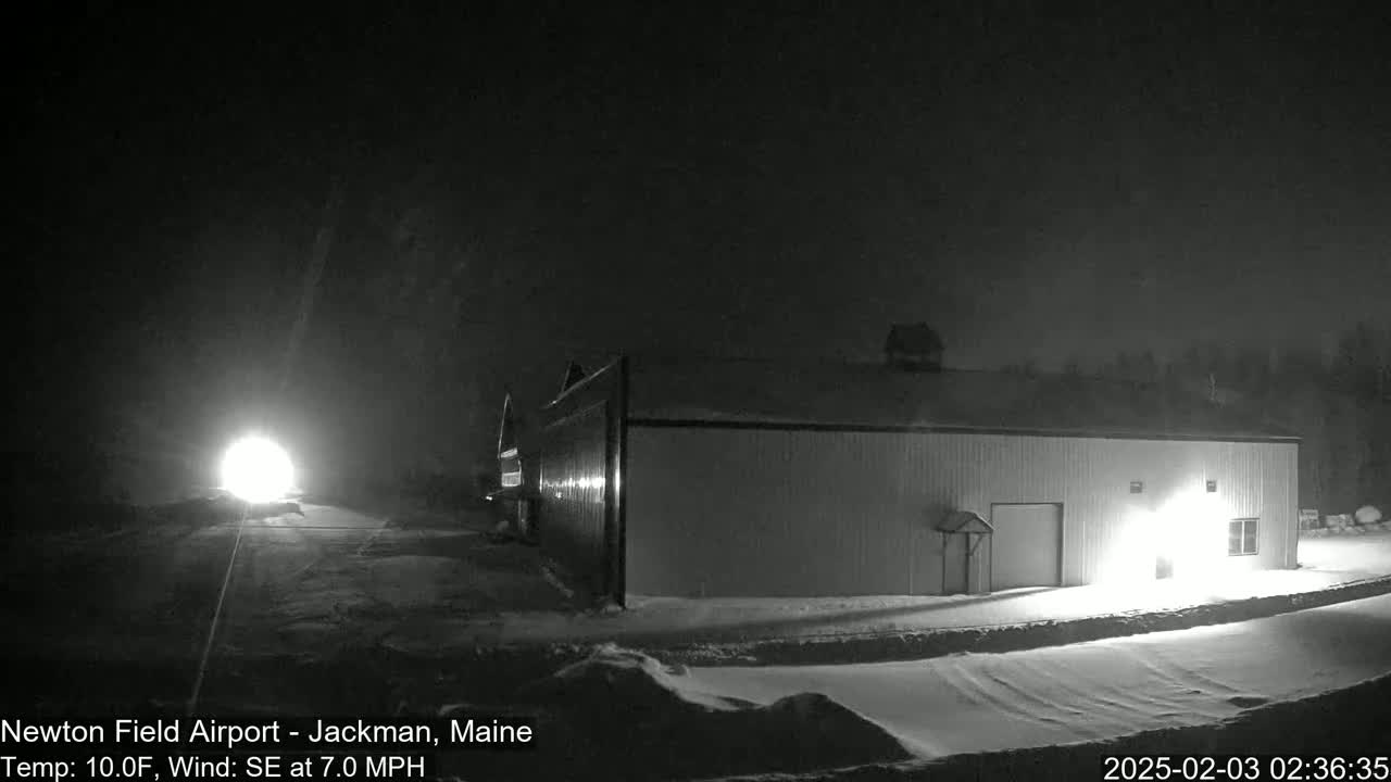 A snow-covered landscape at night shows a long building lit from within, with headlights visible in the distance on a road.