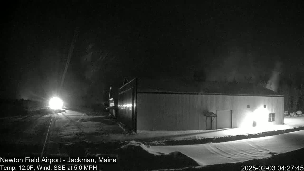 A long, low building is illuminated at night in snowy conditions, with a vehicle's headlights visible in the distance.