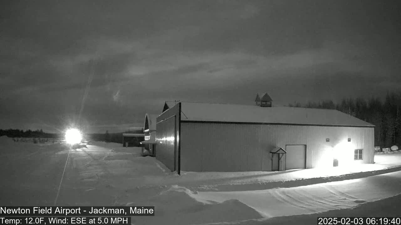 A large hangar and smaller building are illuminated at night in a snowy landscape under a cloudy sky.