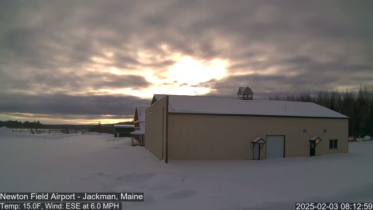 A tan building with a snow-covered roof stands in a snowy landscape under a cloudy sky with the sun peeking through.