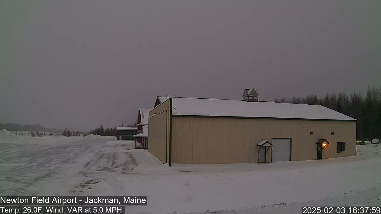 A large tan hangar sits on a snow-covered airfield under an overcast sky.