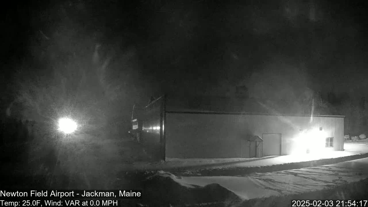 A snow-covered, nighttime scene shows a single-story building illuminated by a bright light, with falling snow visible in the air.