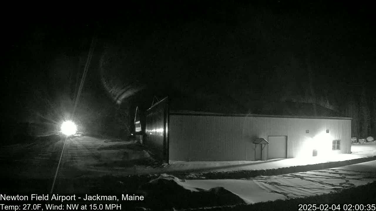 A long, low building is illuminated at night in a snowy landscape under a dark sky with a northwest wind.