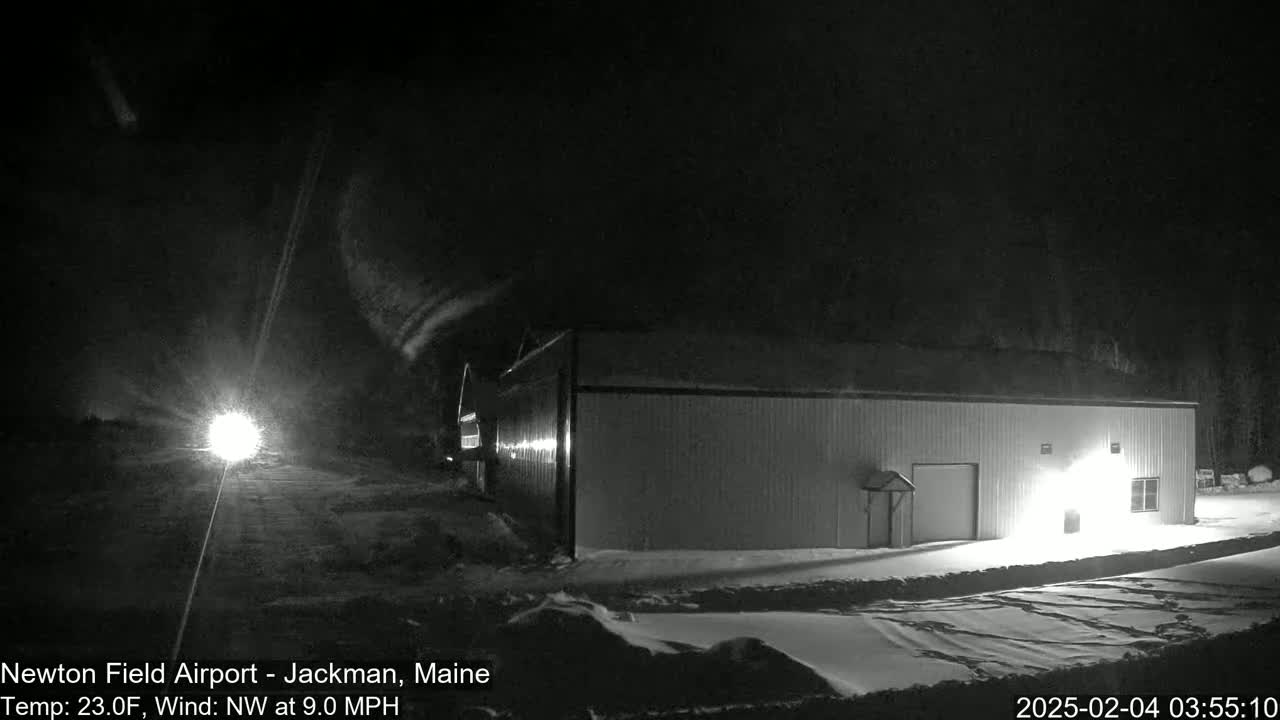 A long, low building is illuminated at night in snowy conditions with a northwest wind.