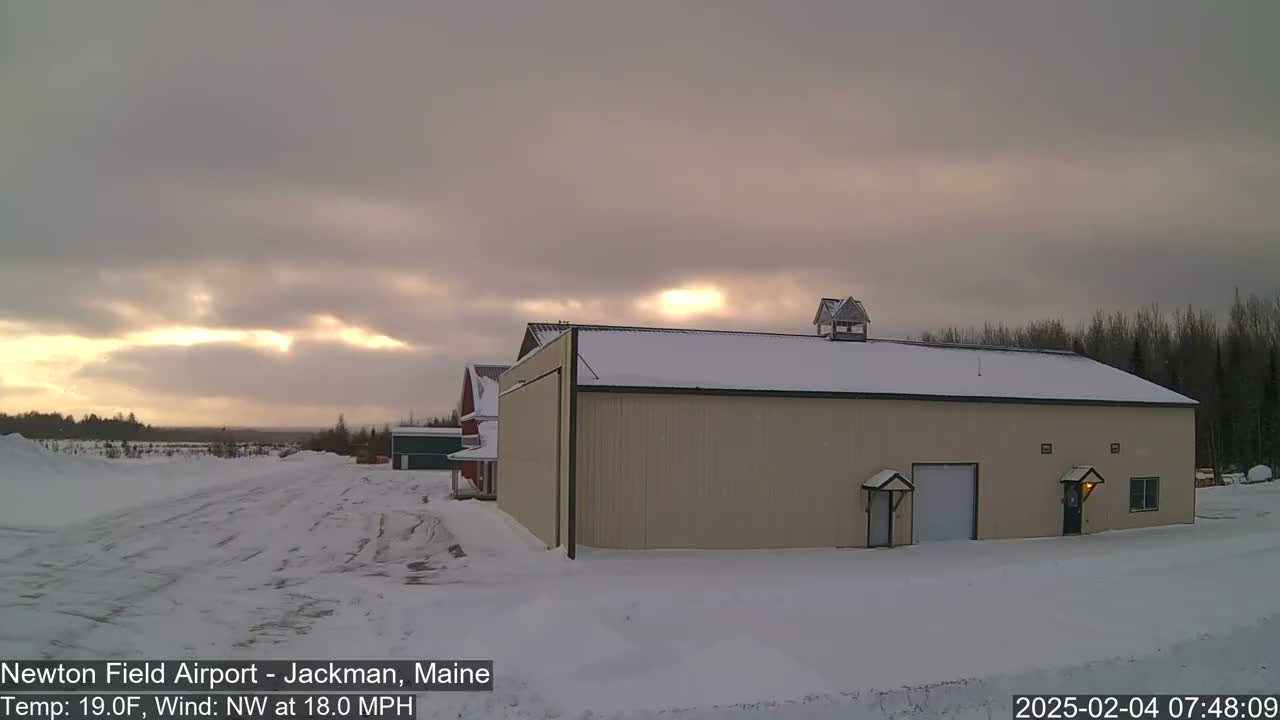A tan hangar sits in a snow-covered landscape under a cloudy, pale sky with a hint of sunrise.