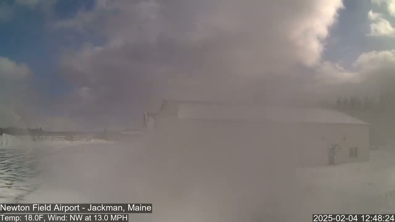 A snow-covered landscape with a building partially obscured by heavy blowing snow under a partly cloudy sky.