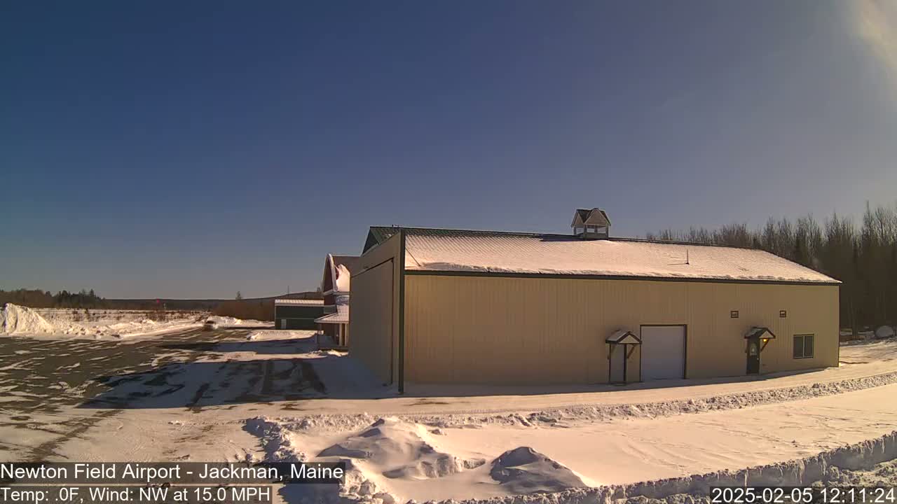 A large tan hangar sits on a snowy airfield under a clear blue sky, with a northwest wind blowing at 15 mph.