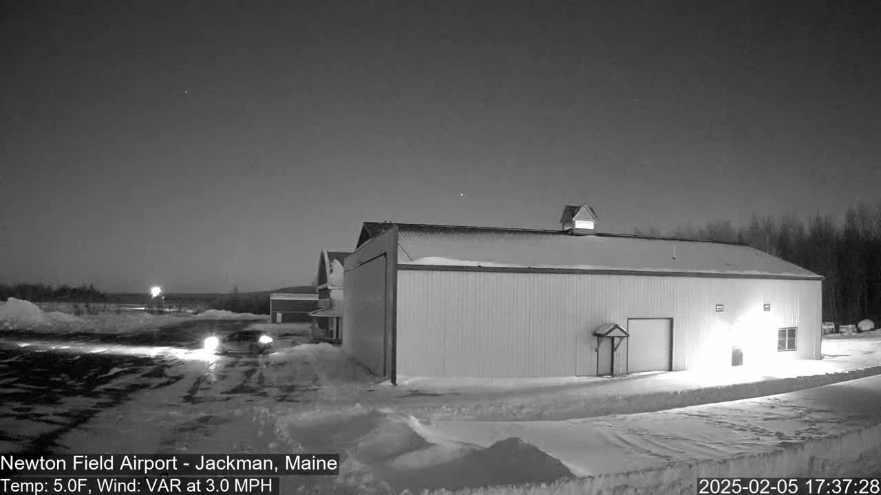 A large, snow-covered hangar sits in a snowy, dimly lit airfield under a dark, clear night sky.