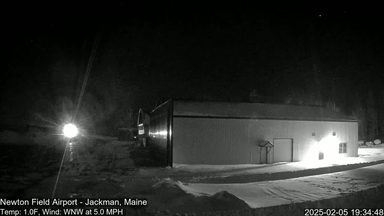 A long, single-story building is illuminated at night in snowy conditions with a northwest wind.