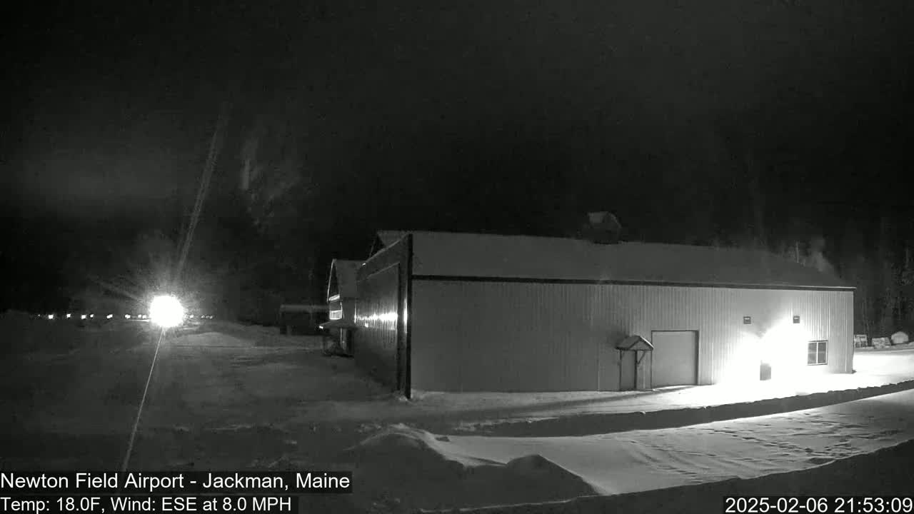 A large, light-colored building sits in a snow-covered area at night under a dark sky with a slight wind.