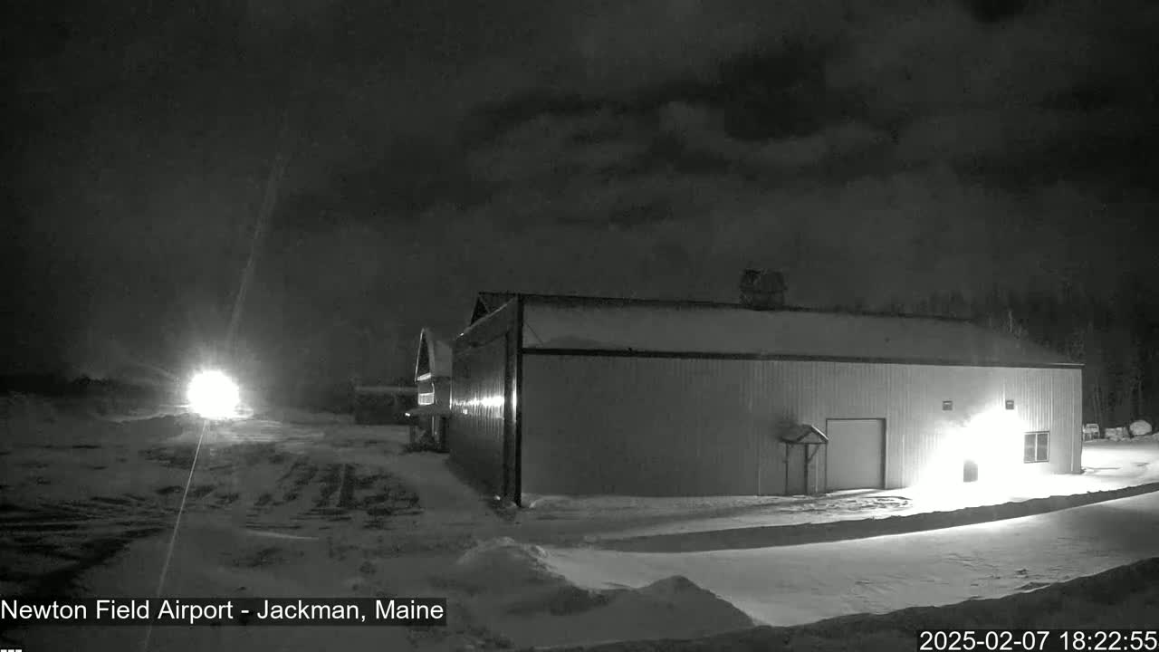 A large, single-story building stands in a snow-covered area at night under a cloudy sky, with bright lights illuminating the scene from a distance.