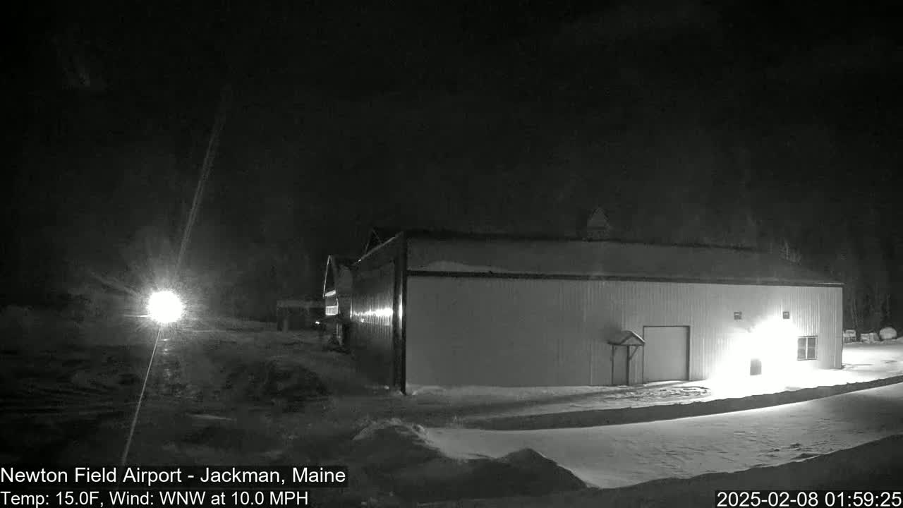 A large, light-colored building is illuminated at night in snowy conditions, with a bright light source visible in the distance under a dark sky.