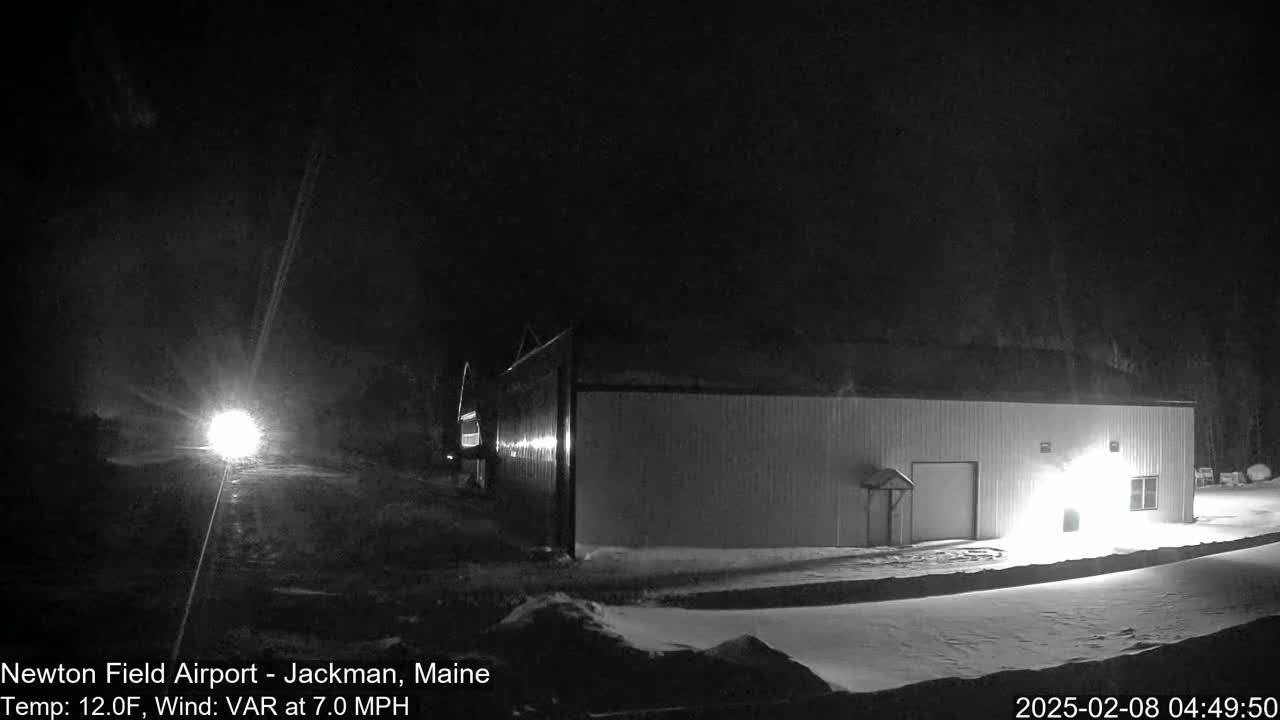 A long, low building is illuminated at night in snowy conditions, with a vehicle's headlights visible in the distance.