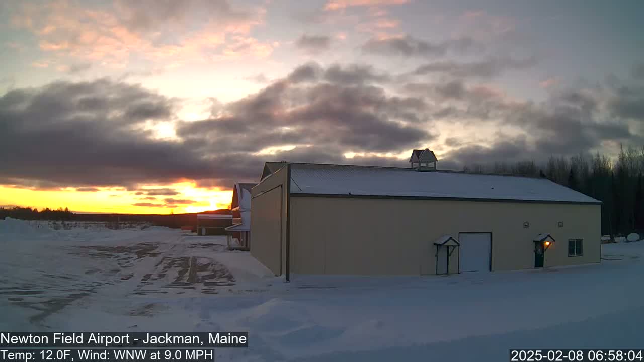 A large tan building with a snow-covered roof sits in a snowy landscape during a sunset with cloudy skies.