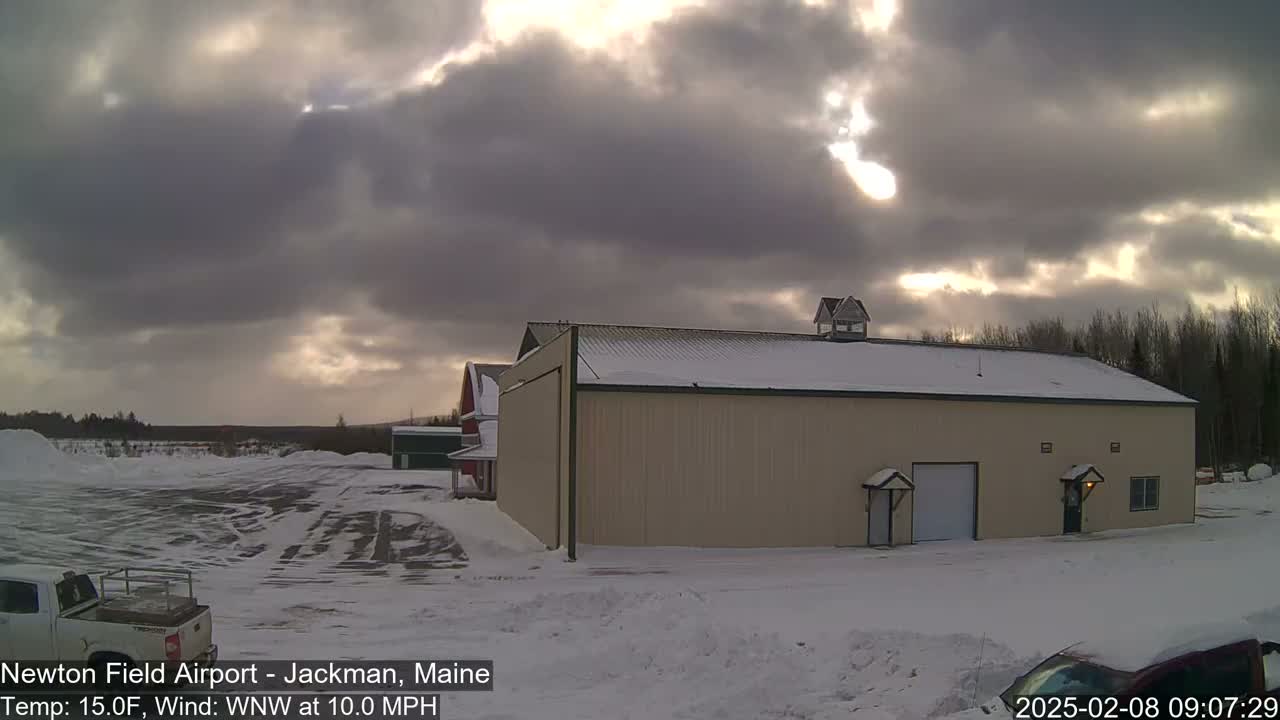 A large tan building with a snow-covered roof sits in a snowy, outdoor area under a partly cloudy sky, with the sun peeking through the clouds.