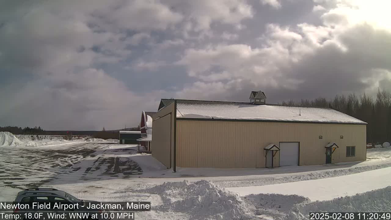 A large tan building with a snow-covered roof sits in a snowy airfield under a partly cloudy sky.