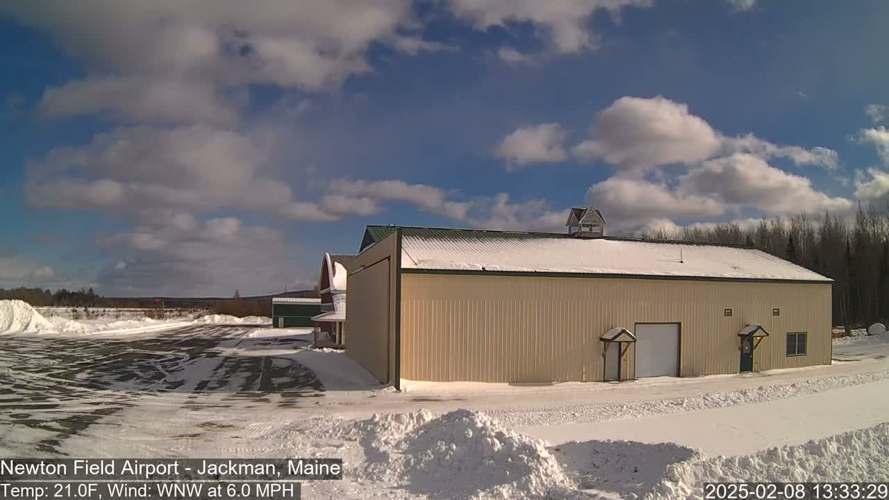 A large tan building with a snow-covered roof sits in a snowy airfield under a partly cloudy sky.