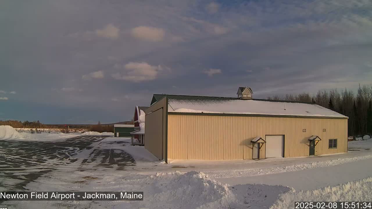 A large tan building with a snow-covered roof sits in a snowy landscape under a partly cloudy sky.