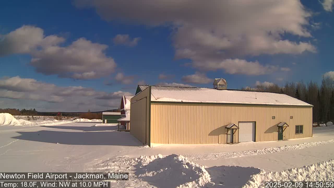 A tan building with a snow-covered roof sits in a snowy landscape under a partly cloudy blue sky.