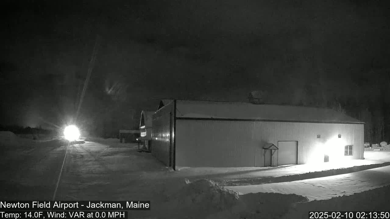 A snow-covered airfield at night, illuminated by a bright light in the distance, features a large, light-colored building to the right.