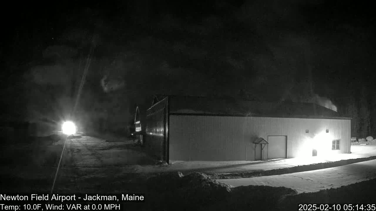 A large, light-colored building is illuminated at night in snowy conditions with calm winds and a temperature of 10 degrees Fahrenheit.