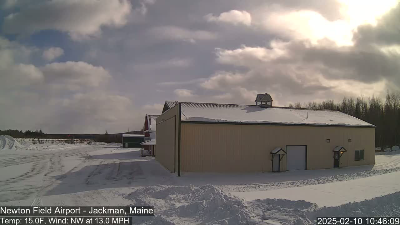 A large tan building with a snow-covered roof sits in a snowy airfield under a partly cloudy sky, with a light wind.