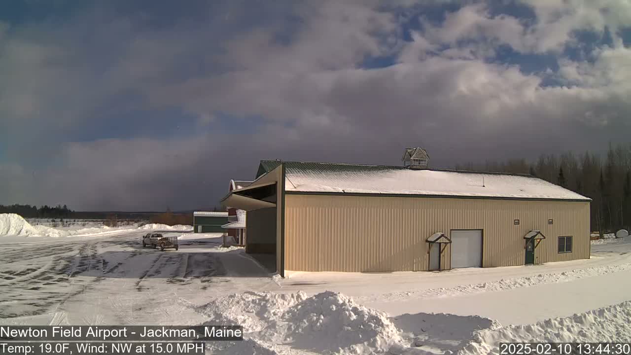 A large tan building with a snow-covered roof sits in a snow-covered airfield under a partly cloudy sky with a northwest wind.